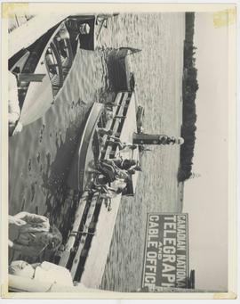 Three motorized watercraft as well as one canoe shown docked at Mooney's Store as many people gat...